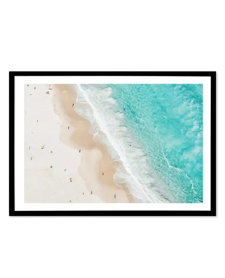 Aerial photograph framed in black showing a coastal beach scene split diagonally. Left side features pale cream sand with small figures of swimmers scattered across it. Center shows white wave foam meeting the shoreline. Right side displays turquoise and aquamarine ocean water with visible depth variation and ripple patterns. Photograph has crisp detail and natural lighting. Black frame with white border.