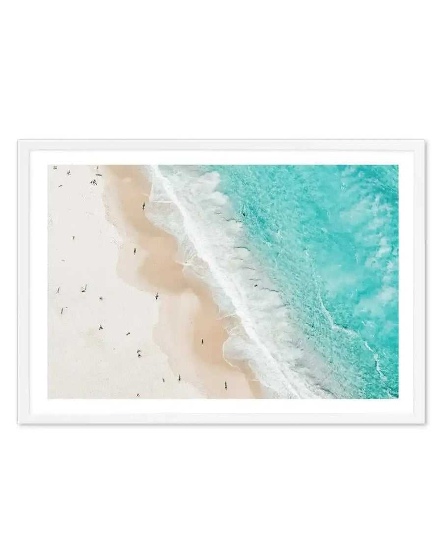 Aerial photograph showing a dramatic coastal scene with a pristine white sandy beach meeting turquoise ocean water. White waves crash along the shoreline while small figures of swimmers and surfers dot the pale sand. The composition transitions from creamy beige sand on the left to crystal-clear turquoise water on the right, with white foam creating a natural dividing line. The photograph is printed in landscape orientation and displayed in a white frame with white border.