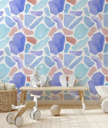 Nursery:
"Peel and stick wallpaper featuring a repeating pattern of abstract, organic shapes in various shades of blue (periwinkle, sky blue, teal), soft lavender, and dusty rose on a cream background. The watercolour design has a loose, painterly quality with visible brushstroke texture. The wallpaper is displayed in a bright nursery above a light wooden bench with a white stuffed bunny, a wooden toy bike, and a light brown ball on the floor."