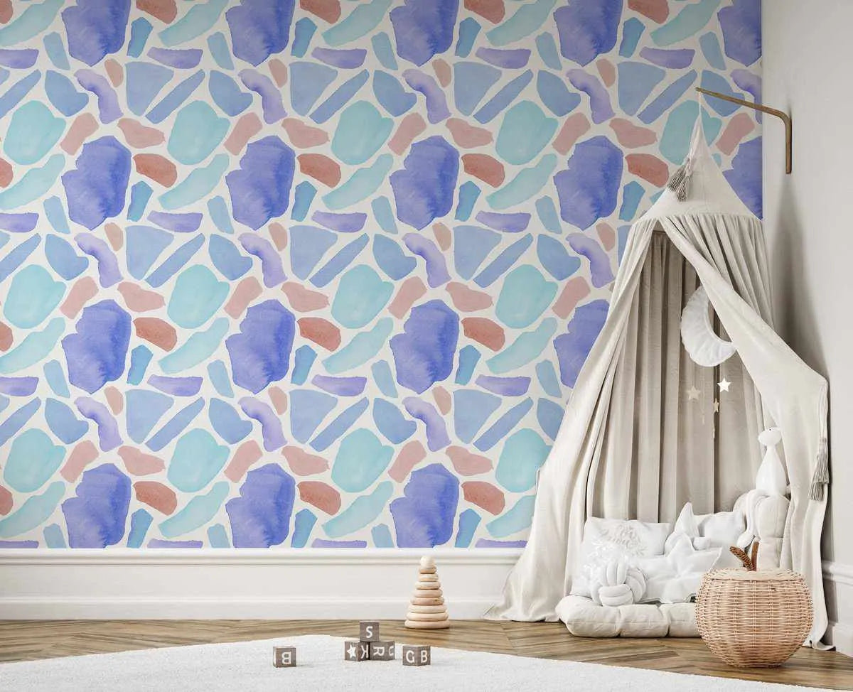 Lifestyle shot of a child's room featuring Bondi Terrazzo Wallpaper. The wallpaper has a repeating pattern of abstract, irregular shapes in shades of light blue, teal, purple, and terracotta, resembling watercolour brushstrokes on a white background. A cream-colored canopy with a round white mobile and star decorations is set up in the corner, creating a cozy nook with white cushions and a woven basket. Wooden alphabet blocks and a stacking toy are on the light wood floor with a white rug.