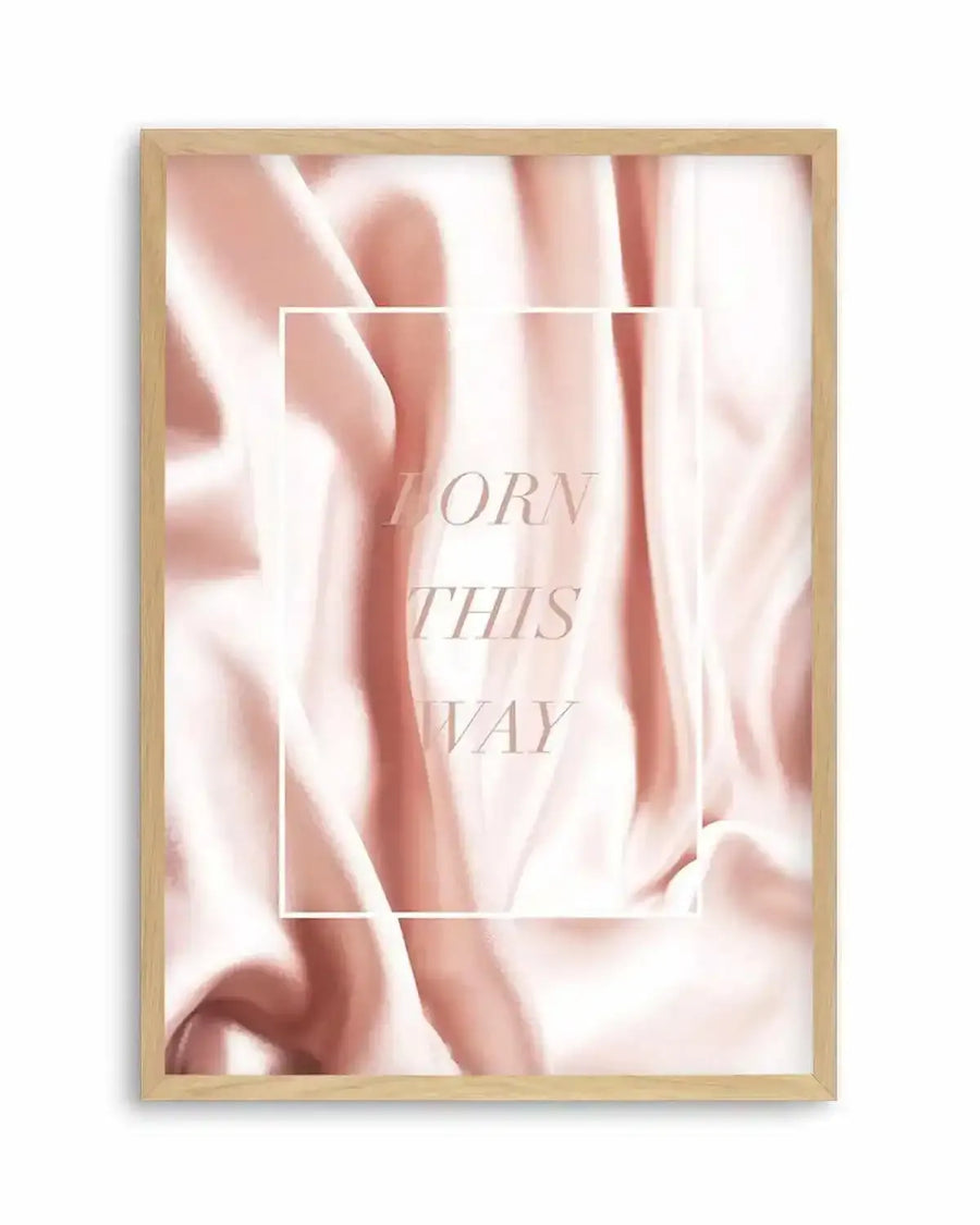 "Framed art print featuring a soft, flowing fabric texture in varying shades of blush pink and cream, creating gentle ripples and folds. The print has a white rectangular overlay in the center with the words 'BORN THIS WAY' in a light, elegant serif font. The print is displayed in a natural oak frame with white border."