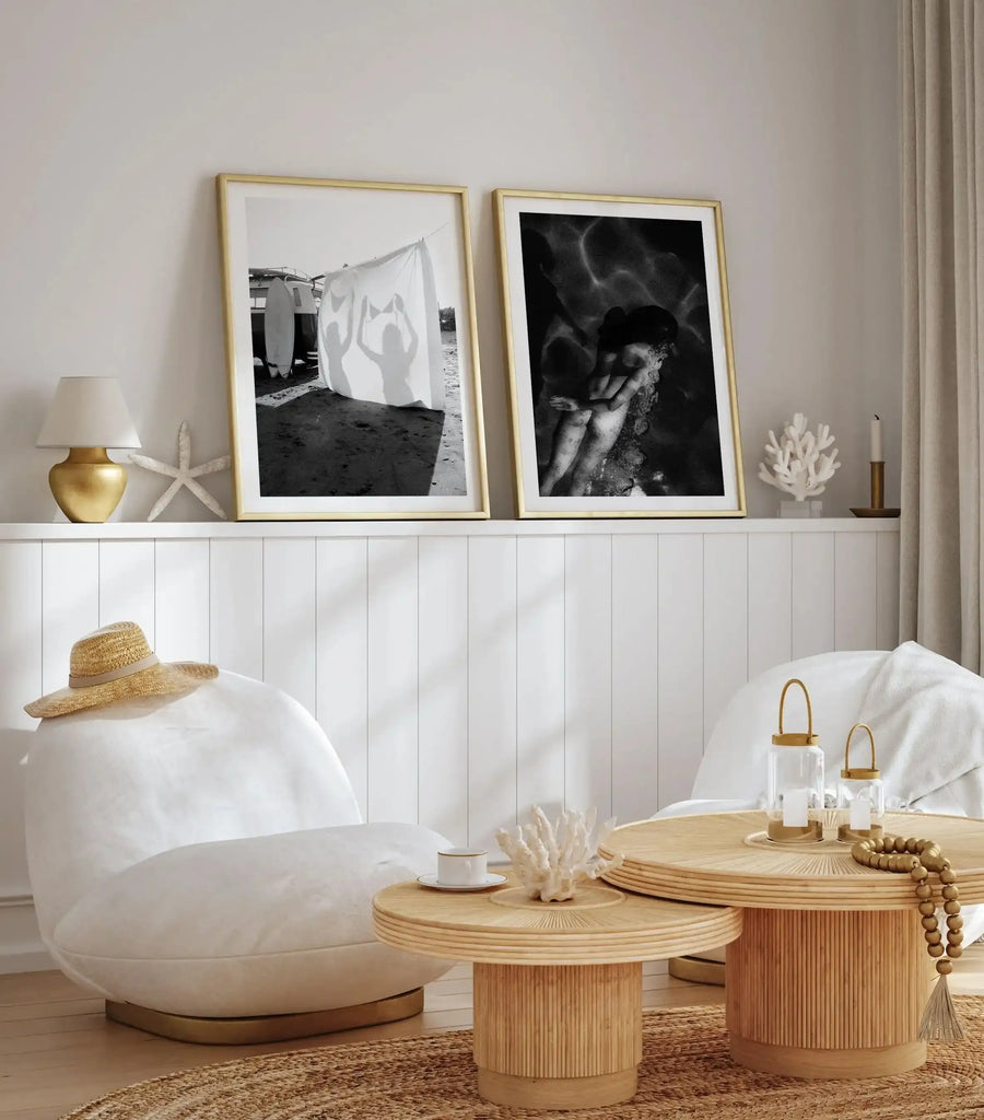 Black and white fine art photograph displayed in a gold-toned frame with white border, hung on a neutral beige wall above white paneled wainscoting. The photograph shows an intimate portrait of a person's face and shoulders in dramatic lighting with strong contrast. Displayed as part of a styled room setting with natural wood side tables, white upholstered furniture, coastal decor including white coral and starfish, brass accents, and neutral textiles.