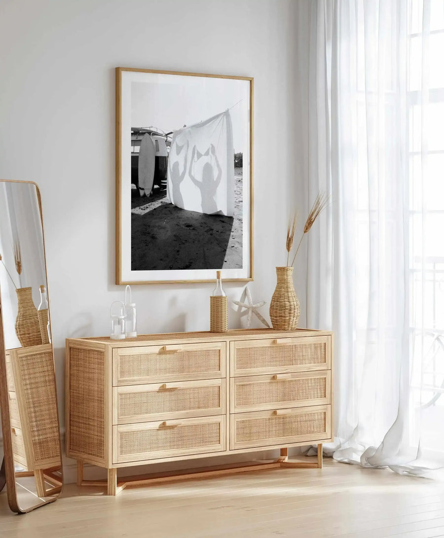 Lifestyle shot of a bedroom featuring a large framed black and white art print above a rattan dresser. The print depicts two silhouetted figures holding up a bikini top against a white sheet on a sandy beach, with a vintage van and surfboards in the background. The room has light wood flooring, a full-length mirror with a natural wood frame, and sheer white curtains over a window. The dresser is adorned with a woven vase, a bottle in a woven holder, and a white starfish.