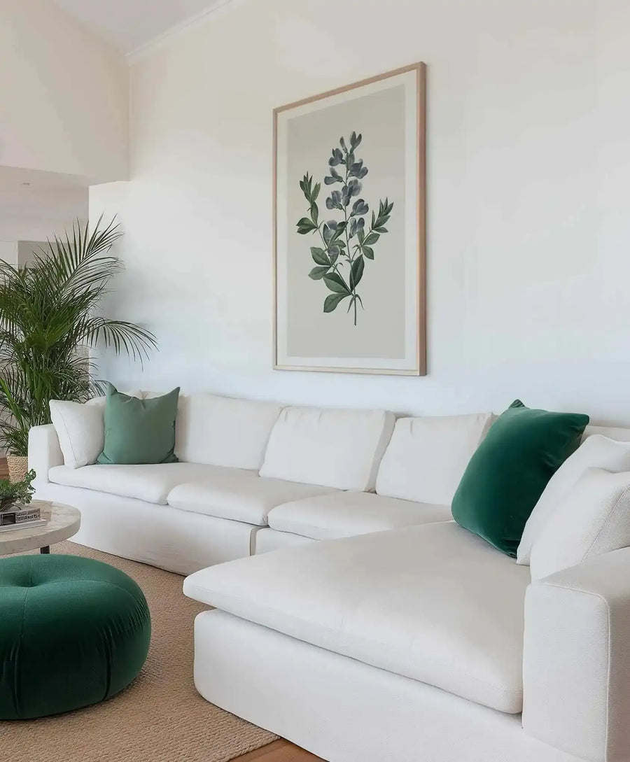 Large framed art print displayed above a white sectional sofa in a bright, minimalist living room. The print features a botanical illustration of a plant with blue-grey leaves and dark green stems on a light cream background, with a natural wood frame and white border. The room includes a large green houseplant, green velvet throw pillows, a round green ottoman, and a round marble coffee table on a woven rug.