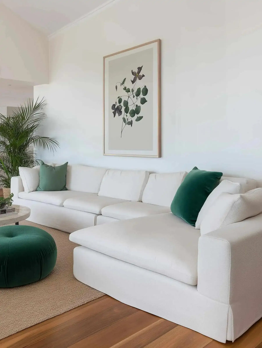 Lifestyle shot of a minimalist living room featuring a large framed art print above a white sectional sofa. The print depicts a botanical illustration of a leafy branch with dark purple flowers and green leaves on a light background, displayed in a natural wood frame with a white border. The room includes green velvet throw pillows and a matching green velvet ottoman, a round marble coffee table, and a large potted plant.