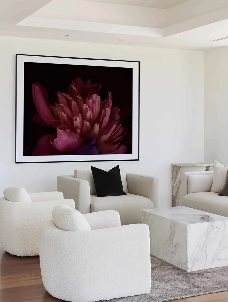 Large framed art print displayed above a curved white sofa in a minimalist living room. The print features a close-up photograph of a deep pink peony flower with layered petals and a dark, moody background, displayed in a black frame with a white border. The room includes two textured cream armchairs, a black velvet cushion, a white marble coffee table, and a light grey rug.