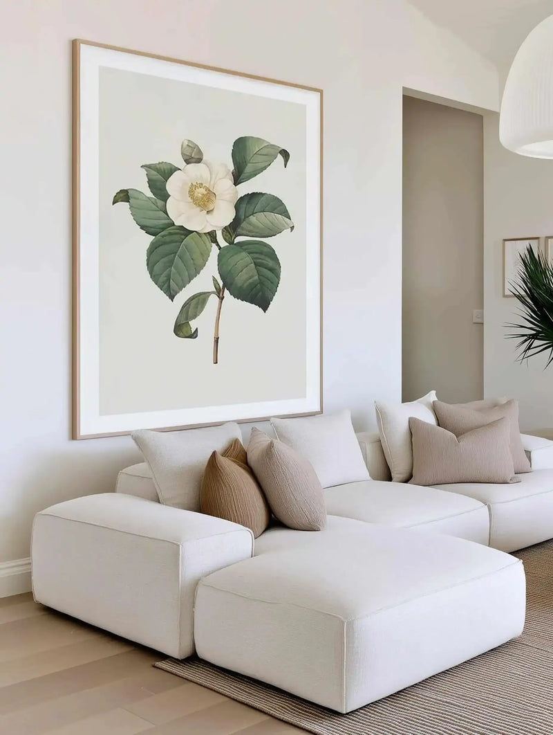 Lifestyle shot of a minimalist living room featuring a large framed art print above a modular white sofa. The print depicts a single white camellia flower with a yellow center and green leaves on a light cream background, displayed in a natural oak frame with a white border. The sofa is adorned with cream and light brown textured throw pillows, and a woven rug lies on the light wood floor.