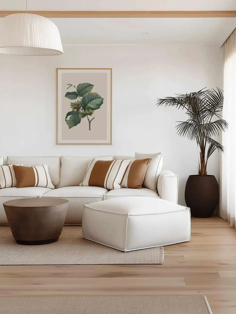Lifestyle shot of a minimalist living room with a large white sofa, a round wooden coffee table, and a white ottoman. Above the sofa, a framed art print features a botanical illustration of a hazelnut branch with green leaves and unripe hazelnuts on a light beige background, displayed in a natural wood frame with a white border. A tall palm plant in a dark brown pot stands in the corner.