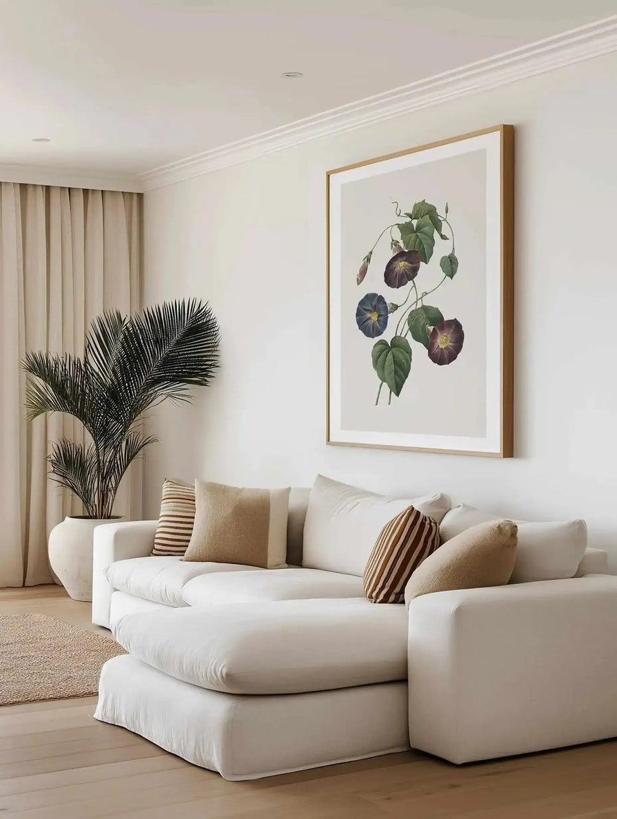 Large framed art print displayed above a white sectional sofa in a bright, minimalist living room. The print features a botanical illustration of morning glory vines with large green leaves and three open flowers in shades of deep purple and blue, with a natural wood frame and white border. The room includes a large potted palm tree in a cream ceramic pot, neutral-toned curtains, and a textured rug.