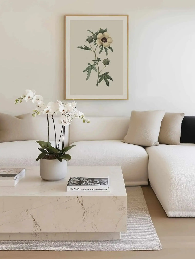 Large framed art print displayed above a white sectional sofa in a minimalist living room. The print features a botanical illustration of a cream-colored flower with a dark brown center and green leaves, set against a light beige background, with a natural wood frame and white border. The room includes a white marble coffee table with books and a white potted orchid.