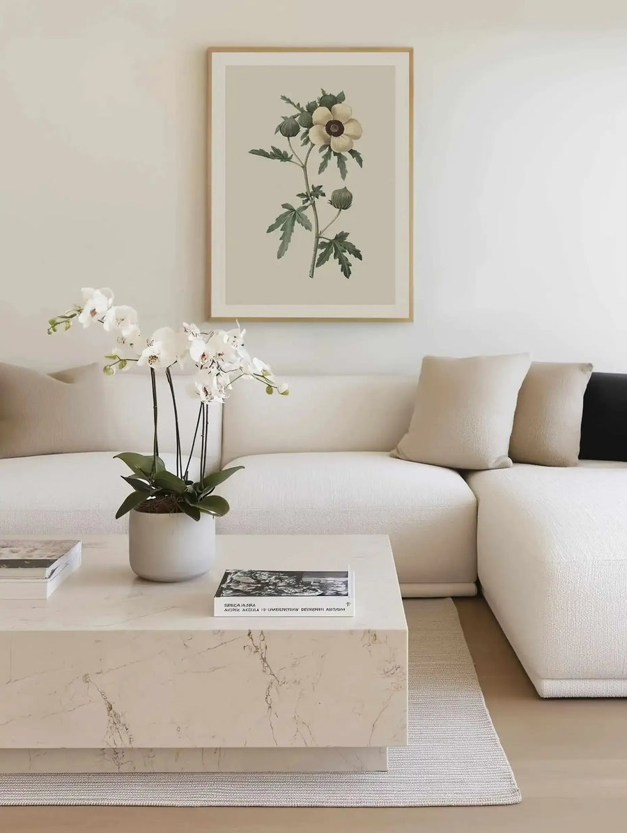 Large framed art print displayed above a white sectional sofa in a minimalist living room. The print features a botanical illustration of a cream-colored flower with a dark brown center and green leaves, set against a light beige background, with a natural wood frame and white border. The room includes a white marble coffee table with books and a white potted orchid.
