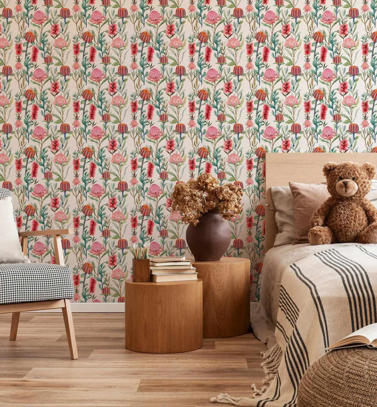 Botanical Garden Wallpaper featuring a repeating pattern of hand-painted Australian native flowers in shades of pink, red, and orange with green foliage on a cream background, displayed on a bedroom wall. The room includes a wooden bed frame, a teddy bear, two round wooden side tables, a dark brown vase with dried flowers, and a black and white houndstooth armchair.