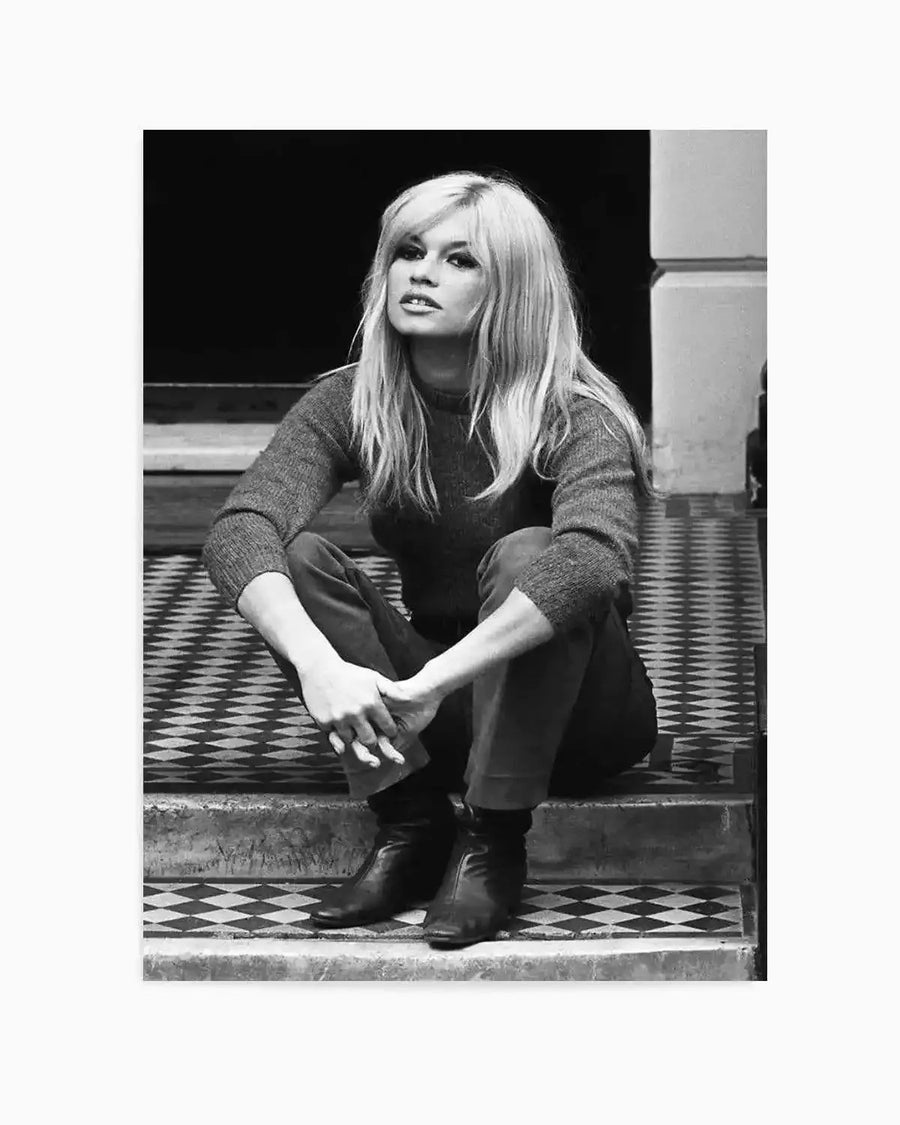 Black and white photographic art print of Brigitte Bardot sitting on steps. The composition features a full-length shot of the blonde actress with long hair and bangs, wearing a dark sweater and pants with black boots, looking directly at the viewer. She is seated on steps with a diamond-patterned tile floor.