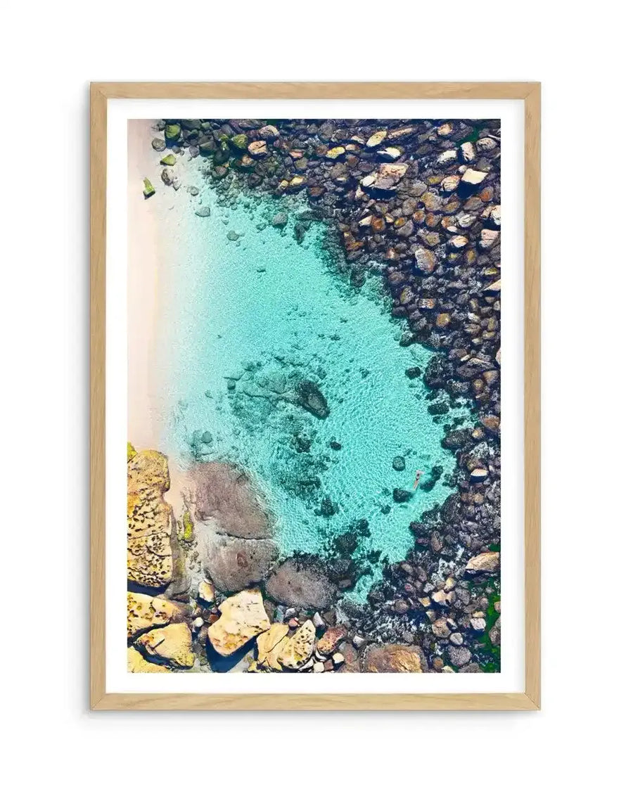 Framed aerial photography art print of Bronte Beach Pool with a natural wood frame and white border. The overhead shot captures a turquoise swimming pool carved into a rocky coastline, with a small strip of white sand on the left and a lone swimmer in the pool.