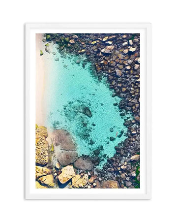 Aerial photograph of Bronte Beach Pool framed in white with white border, showing crystalline turquoise water surrounded by dark basalt rocks and golden-yellow lichen-covered boulders. The composition captures the pool's shallow turquoise depths with scattered dark rocks visible beneath the surface, contrasting with the darker rocky coastline framing the edges. The image conveys a serene coastal landscape in natural daylight.