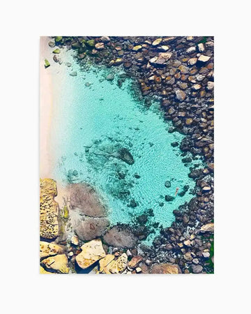 Aerial photograph of Bronte Beach Pool showing turquoise water surrounded by dark rocky outcrops and golden-yellow lichen-covered boulders. The crystalline water displays depth gradients from pale aqua near the sandy bottom to deeper teal, with visible rocks scattered throughout. Rocky shoreline frames the composition on all sides, captured from directly above. Unframed art print.
