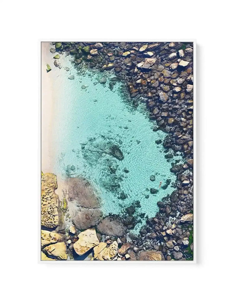 Gallery-wrapped canvas print of an aerial photograph of Bronte Beach Pool, featuring clear turquoise water surrounded by a sandy beach on the left and dark, textured rocks on the right. A small figure is visible swimming in the pool. The composition captures the vibrant colours and natural textures of the coastal scene.