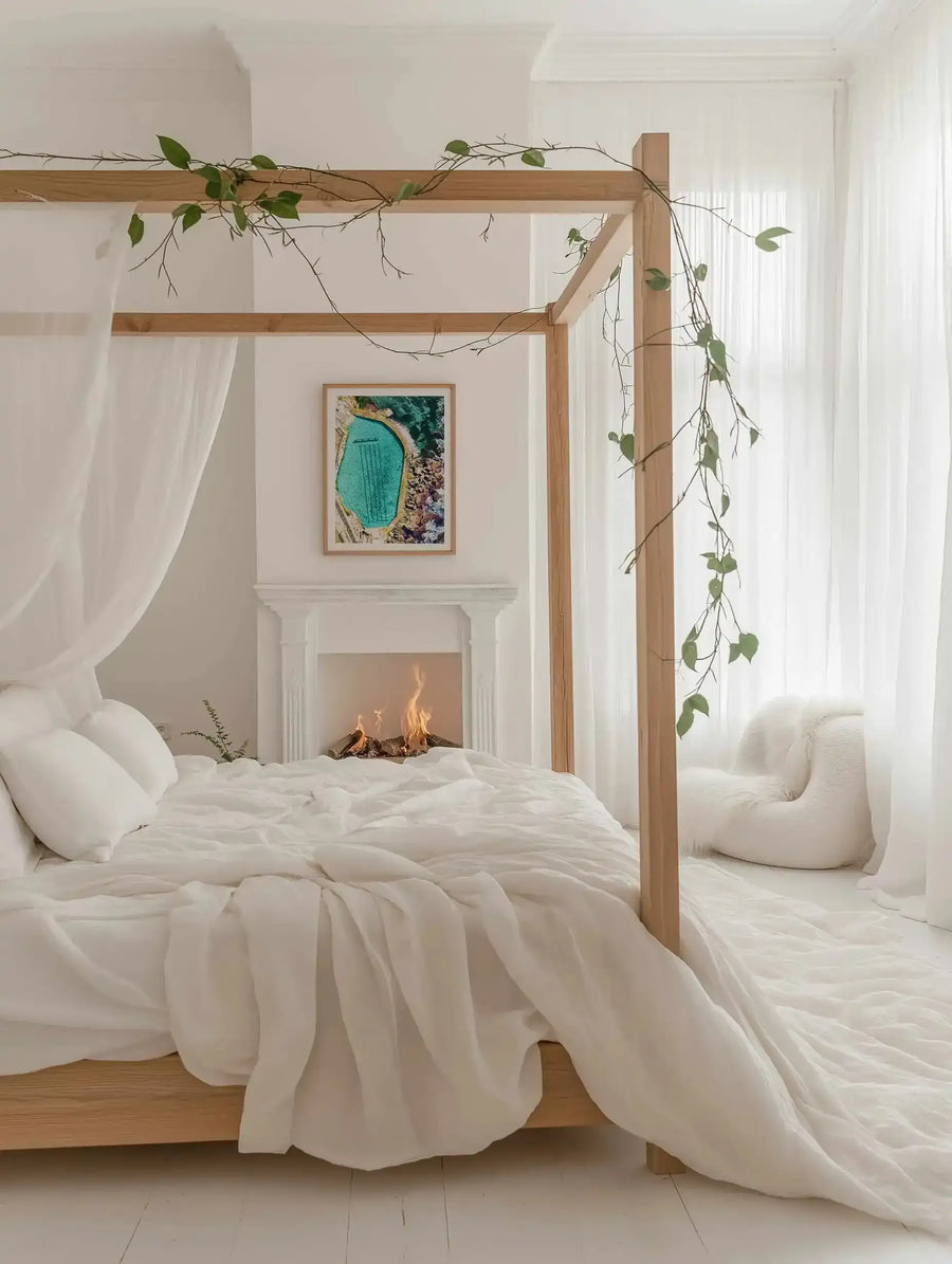 Lifestyle shot of a minimalist bedroom with a framed art print displayed above a white fireplace with a lit fire. The art print features an aerial photograph of the Bronte Ocean Pool, a turquoise swimming pool surrounded by rocks and ocean, with a natural wood frame and white border. The room includes a wooden four-poster bed with white linen bedding and sheer white curtains, and a white fluffy armchair in the corner.