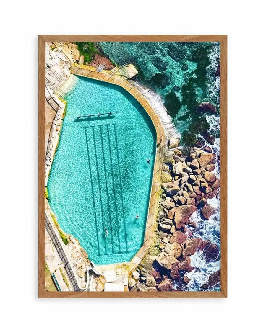 Framed aerial photograph of the Bronte Ocean Pool in Sydney, Australia, with a natural wood frame and white border. The vibrant turquoise pool, divided into swimming lanes, is nestled against a rocky coastline where clear blue ocean water meets the shore with gentle white waves. Swimmers are visible in the pool.