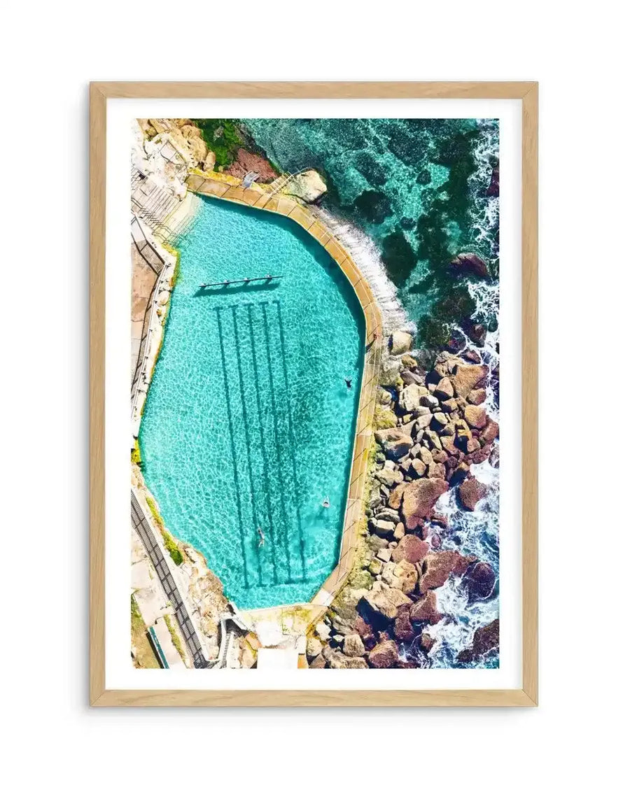 Framed aerial photograph of the Bronte Ocean Pool in Sydney, Australia, with a natural wood frame and white border. The composition captures the bright turquoise water of the rectangular pool with four dark lane lines, surrounded by a concrete edge and rugged brown rocks. Beyond the pool, the clear blue ocean with white waves crashes against the shore.