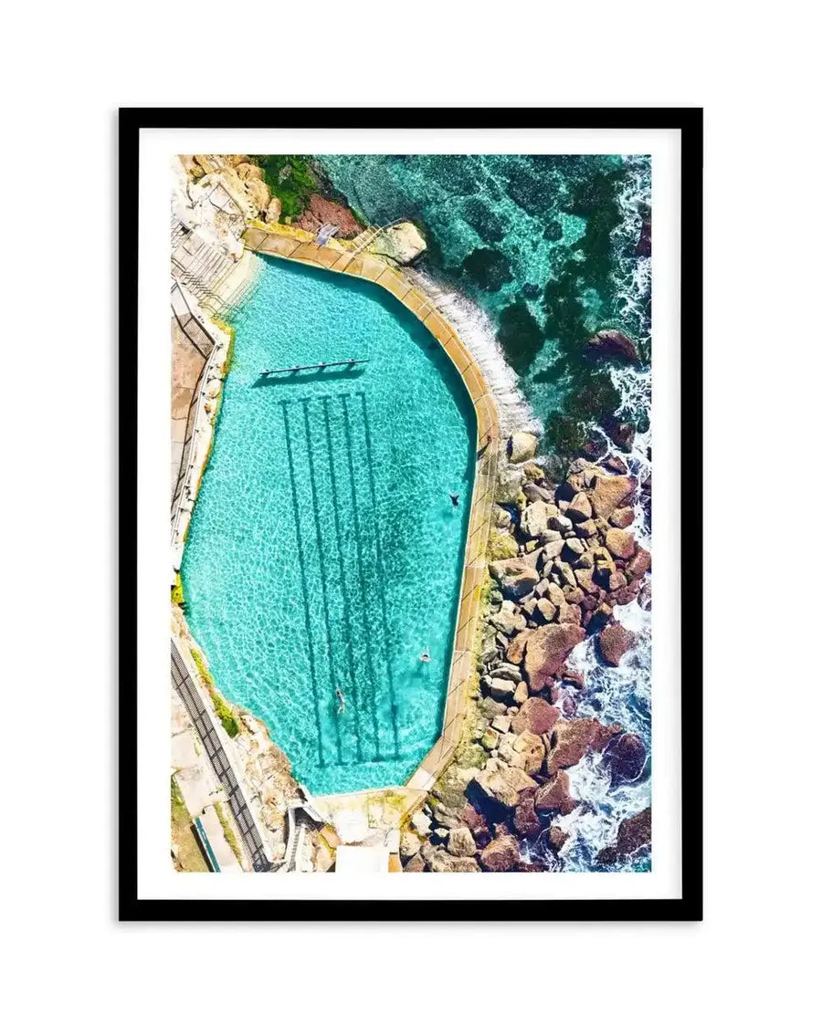 Aerial photograph of Bronte Ocean Pool framed in black with white border, showing a turquoise oval-shaped saltwater pool enclosed by golden sandstone walls and timber decking on the left side, with dark green ocean, white foaming waves, and reddish-brown rocky outcrops visible on the right edge, taken from directly above.