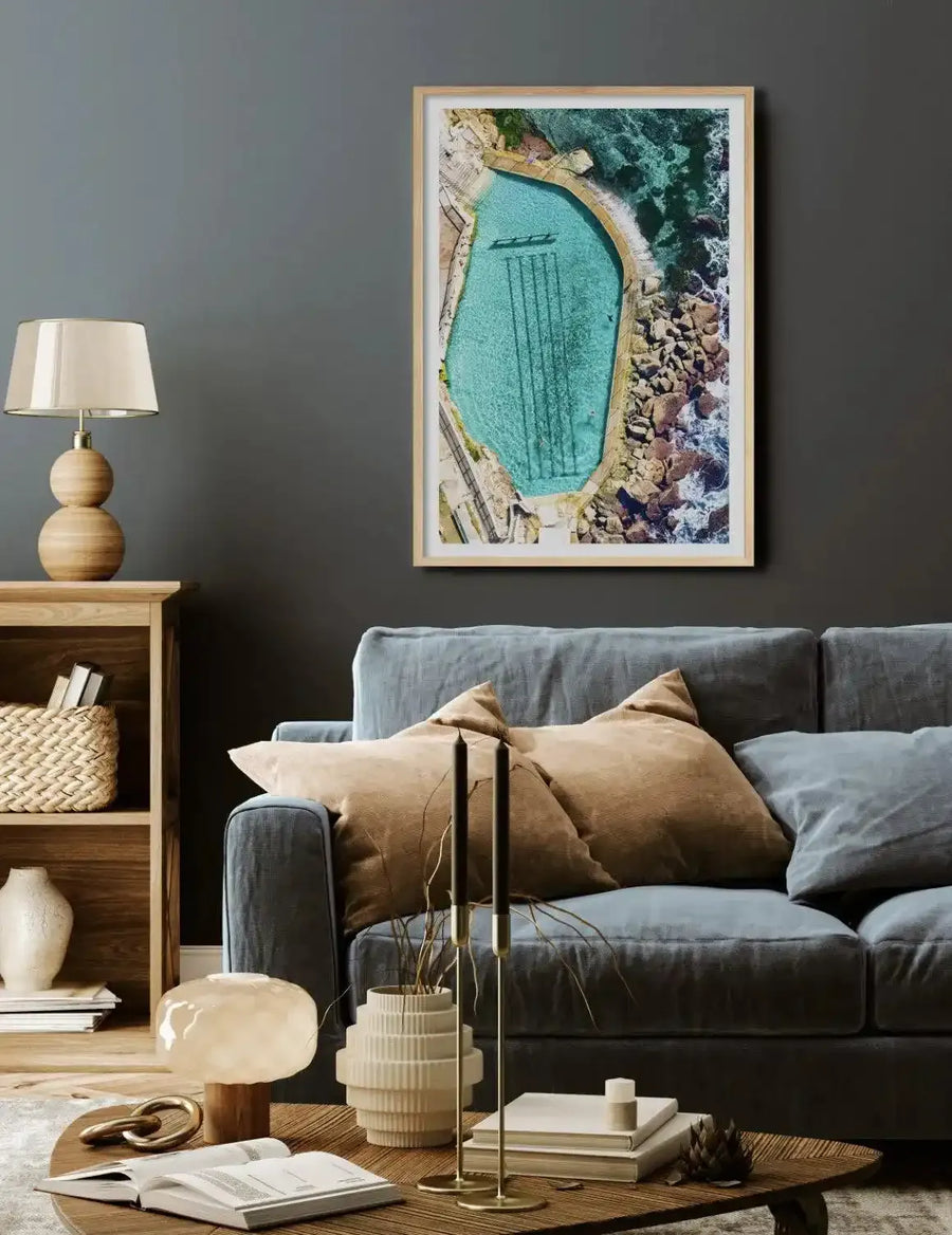 Aerial photograph of Bronte Ocean Pool framed in light wood with white border, displaying an oval-shaped saltwater pool with turquoise water surrounded by rocky coastal cliffs, golden sand, and dark ocean waves. The framed print hangs on a dark grey wall above a grey linen sofa styled with tan and grey cushions. Natural wood side table with lamp, woven basket, and decorative objects visible in foreground.