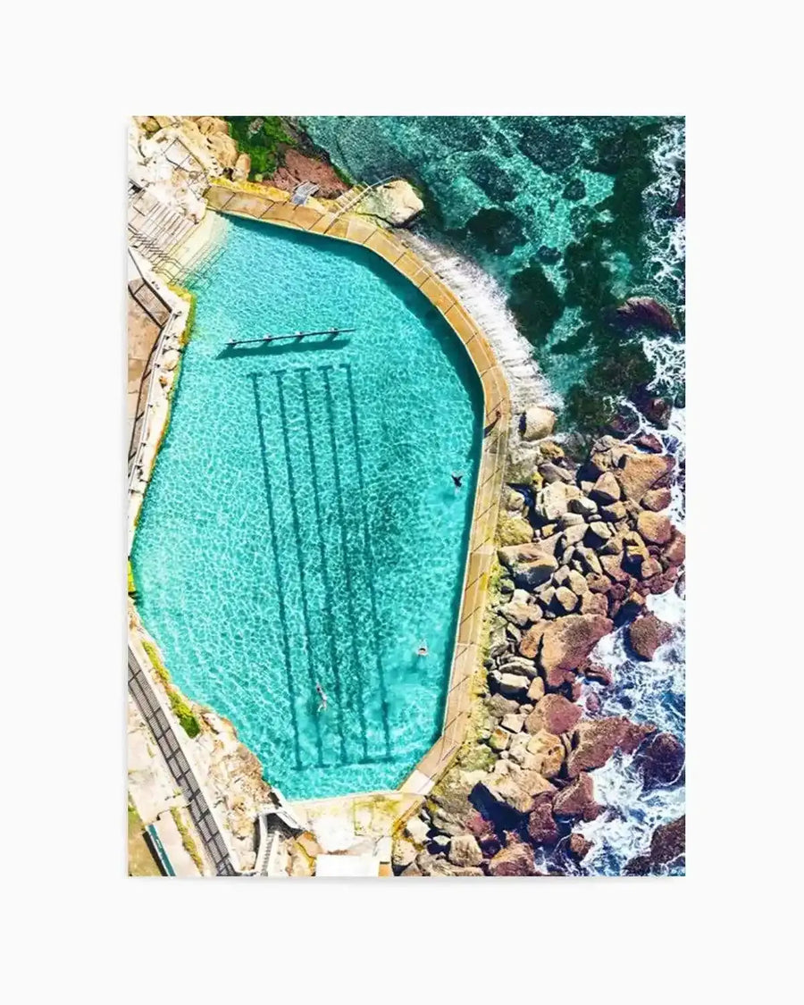 Aerial photograph of Bronte Ocean Pool in Sydney, showing a curved turquoise swimming pool with yellow safety railings enclosed by rocky breakwalls. Pristine cyan water contrasts with dark teal ocean, white wave foam, and rust-coloured boulders. The composition captures the pool's distinctive shape and surrounding coastal landscape in vibrant natural colours, presented as an unframed art print.