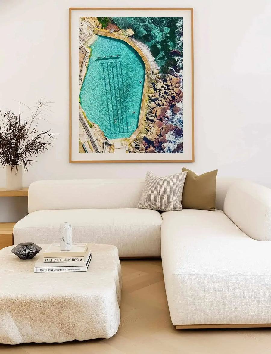 Framed aerial photograph of Bronte Ocean Pool displayed above a curved cream linen sectional sofa in a minimalist living room. The artwork shows a turquoise swimming pool bordered by golden-tan concrete, surrounded by rocky dark teal ocean waters and white wave foam. The photograph is mounted in a natural wood frame with white border. Two textured throw pillows in cream and olive tones rest on the sofa. A curved cream ottoman and potted plant with thin green foliage flank the seating arrangement.