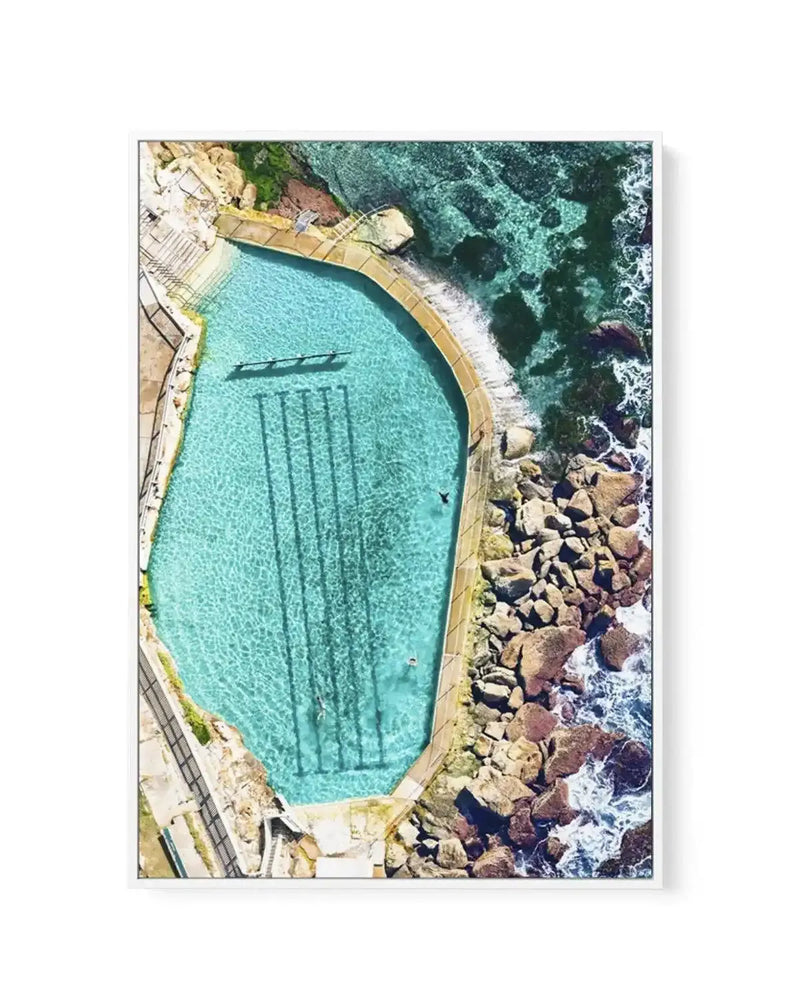 Gallery-wrapped canvas print of an aerial photograph of the Bronte Ocean Pool in Sydney, Australia. The image captures the turquoise blue water of the man-made pool, with four dark lane lines visible on the bottom, and several swimmers. The pool is surrounded by a concrete edge and natural rock formations, with the darker blue ocean and white waves crashing against the rocks on the right side of the frame. The composition is bright and vibrant, showcasing the clear water and rugged coastline.