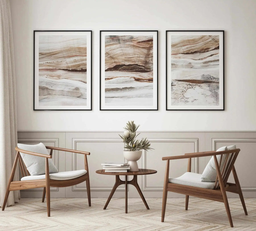 Lifestyle shot of a minimalist living room featuring three framed abstract art prints displayed above a wainscoting wall. The prints feature horizontal layers of natural rock formations in shades of beige, brown, and white, with visible striations and textures, framed in black with a white border. Two light wood armchairs with white cushions flank a small round wooden table with books and a white vase with dried foliage.