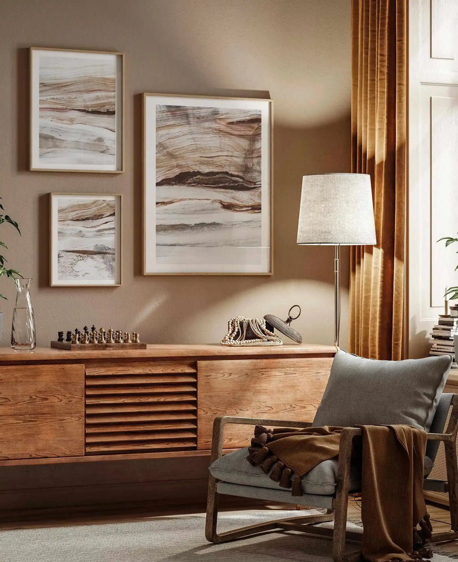 Three framed art prints displayed on a warm beige wall above a wooden console table in a cozy living room. The prints feature abstract, layered compositions with horizontal bands of earthy brown, cream, and grey, suggestive of natural rock formations or stratified landscapes, with natural wood frames and white borders. The room includes a grey armchair with a brown throw, a floor lamp, a chess set, and a string of pearls on the console table.