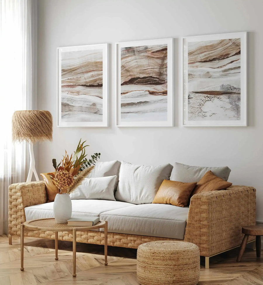 Lifestyle shot of three framed art prints displayed above a woven rattan sofa in a bright living room. Each print features a close-up photograph of layered rock formations with horizontal bands of light cream, warm beige, and deep reddish-brown, suggestive of sandstone or marble. The prints are displayed in simple white frames with white borders. The room includes a large window with sheer white curtains, a woven floor lamp, a small round coffee table, and a woven jute ottoman.