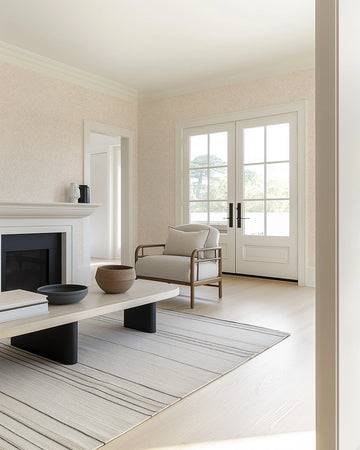 "Bronte Wallpaper featuring a repeating pattern of small, hand-drawn, radiating dashes in a soft, muted blush pink on a light cream background, covering the walls of a bright living room. The wallpaper is visible above a white fireplace mantel and around a large doorway, complementing light wood floors, a striped rug, and a light-colored armchair."