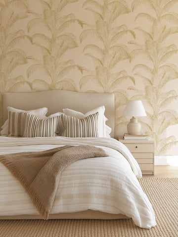 Lifestyle shot of a bedroom featuring Bronzed Palms wallpaper. The wallpaper has a repeating pattern of hand-drawn palm fronds in a bronzed gold hue on a warm cream background, creating a tropical yet sophisticated ambiance. The wallpaper is visible behind a bed with a cream upholstered headboard, white bedding with subtle stripes, and striped throw pillows in cream and brown. A textured brown throw blanket is draped over the bed. A cream nightstand with a ceramic lamp and books sits on the right.