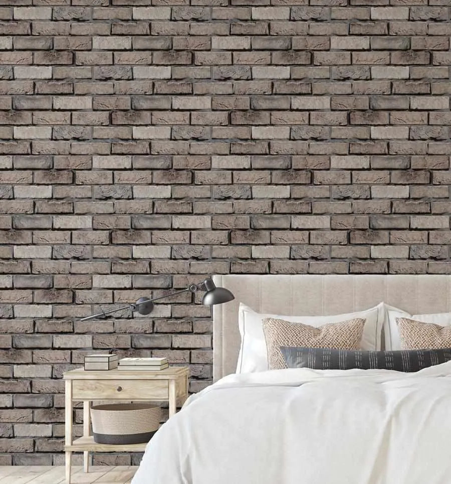 Brown brick wallpaper with a repeating pattern of rectangular bricks in varying shades of brown, grey, and charcoal, creating a textured and rustic look. The wallpaper covers the wall behind a bed with a cream upholstered headboard, white duvet, and decorative pillows in neutral tones. A light wood bedside table with books and a woven basket sits to the left, and a black wall-mounted lamp extends over the bed.