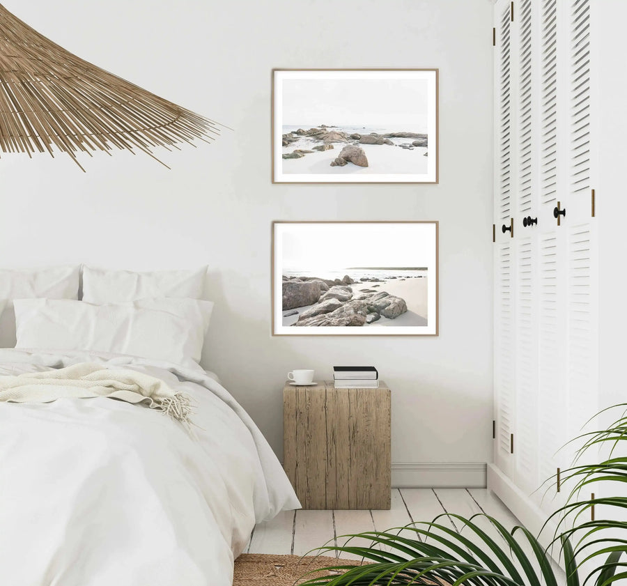 Lifestyle shot of a minimalist bedroom featuring two framed art prints above a wooden bedside table. The prints are photographs of a sandy beach with large, smooth rocks and gentle waves under a bright, overcast sky, displayed in natural wood frames with white borders. The room has white walls, a bed with white linen and a cream textured throw, a white slatted wardrobe with brass handles, and a green plant in the foreground.
