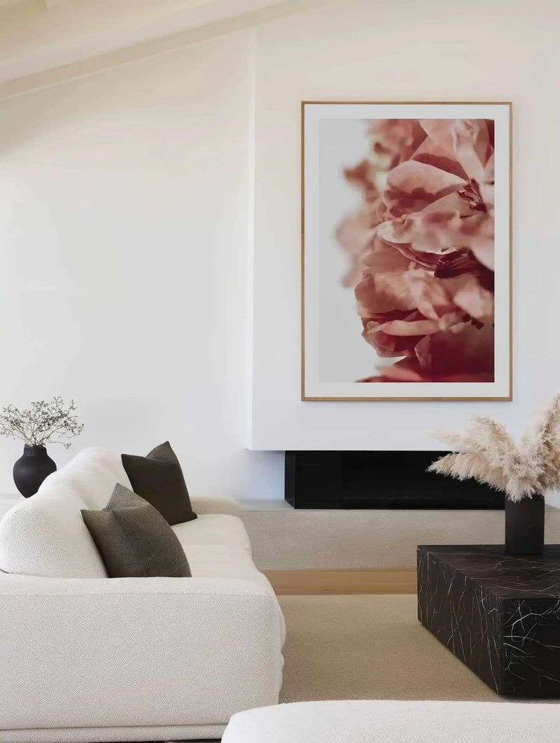 Living room: Large framed art print displayed above a modern fireplace in a minimalist living room. The print features a close-up photograph of a burnt orange peony with soft, textured petals and a white background, displayed in a natural oak frame with white border. The room includes a textured white sofa with dark grey cushions, a black marble coffee table, and dried pampas grass in a black vase.