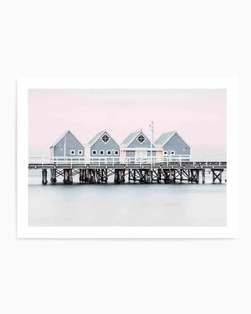 Framed photograph of the Busselton Jetty in Western Australia, featuring a row of four small, light grey wooden beach houses with white trim and white picket fences, standing on a long wooden pier over calm, pale blue water. The sky above is a soft, pastel pink, creating a serene and minimalist coastal scene.