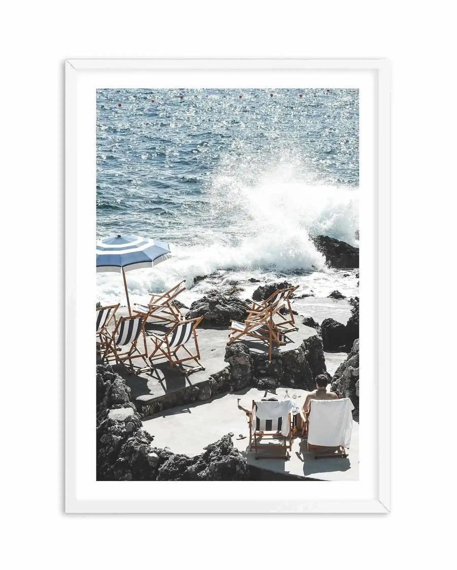 Framed photography art print of a rocky beach in Capri with a white frame and white border. The scene features a blue and white striped umbrella and several empty wooden deck chairs with striped fabric on a concrete platform surrounded by dark rocks. In the foreground, a person sits in a white deck chair with their back to the viewer, facing the ocean where a large wave crashes against the rocks. The water is a sparkling blue with small red buoys visible in the distance.