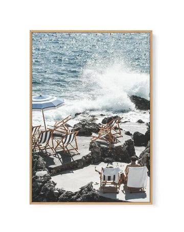 Framed canvas art print featuring a coastal beach scene in natural wood frame. The photograph captures a rocky Capri shoreline with crashing waves, several striped beach chairs and loungers arranged on the sand, a blue and white striped beach umbrella, and people relaxing by the turquoise water. Rocky dark outcroppings frame the composition. The image has a bright, sunny Mediterranean aesthetic with white borders on the canvas.