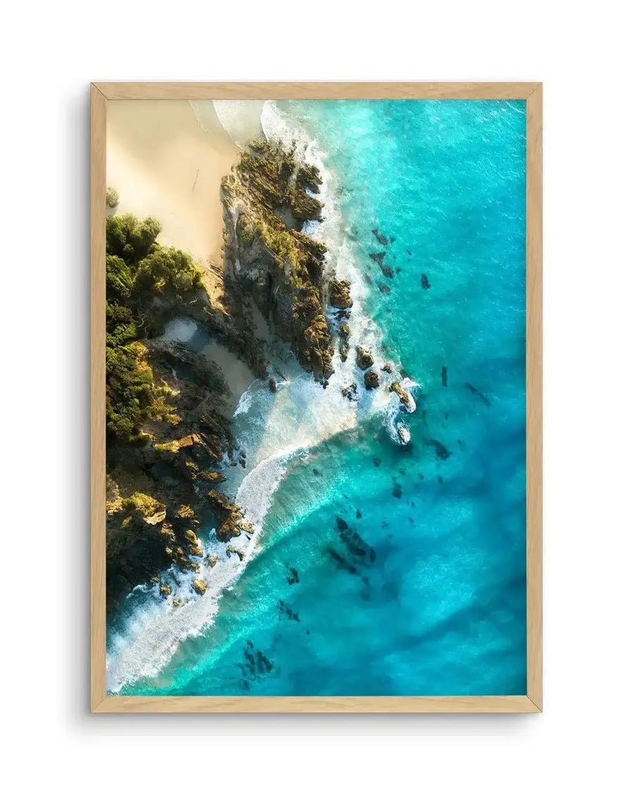Aerial photography art print of a coastal scene with vibrant turquoise ocean water meeting a sandy beach and rocky shoreline, displayed in a natural oak frame with white border. Waves crash against the rocks and sand, creating white foam, and lush green vegetation grows on the land.