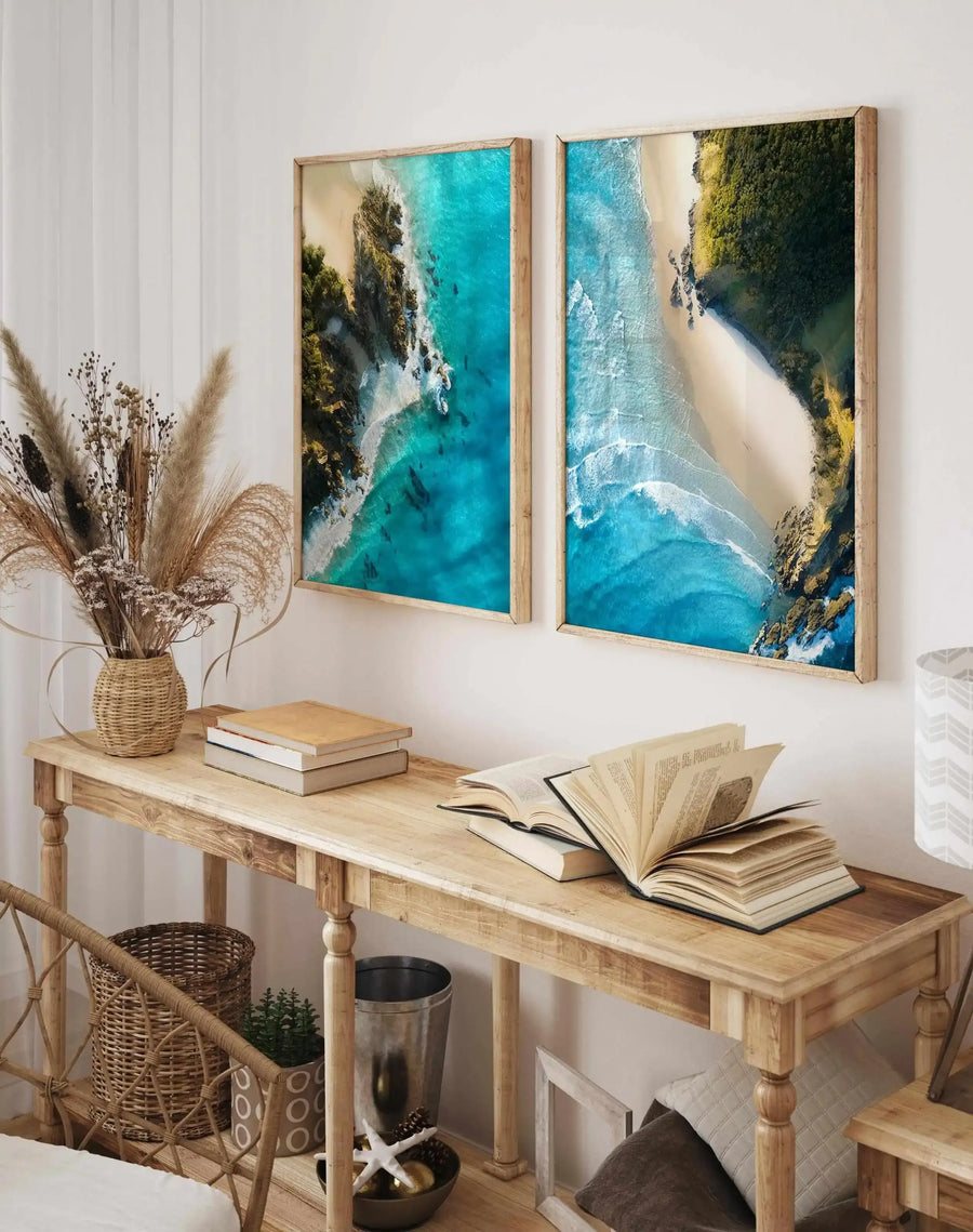 Lifestyle shot of two framed art prints displayed above a wooden console table in a neutral-toned room. The prints feature aerial photography of a tropical beach with turquoise water, white sand, and lush green vegetation, displayed in natural wood frames with white borders. The console table holds a woven vase with dried pampas grass, stacks of books, and a table lamp with a white chevron pattern shade.