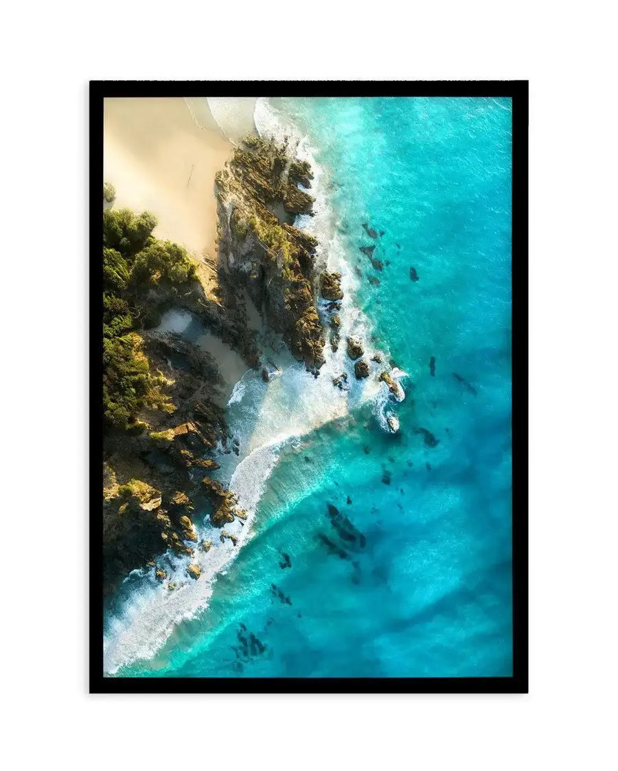 Framed aerial photograph of a coastal beach scene with a black frame and white border. The composition features a sandy beach meeting a rocky coastline, with vibrant turquoise ocean water crashing against the shore, creating white foamy waves.