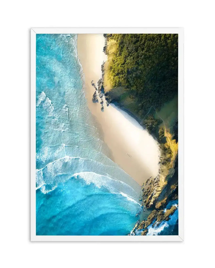 Framed aerial photograph of a coastal beach scene with a white frame and white border. The composition features a long stretch of golden sand bordered by turquoise ocean waves on the left and lush green forest on the right, with rocky outcrops visible where the forest meets the beach.