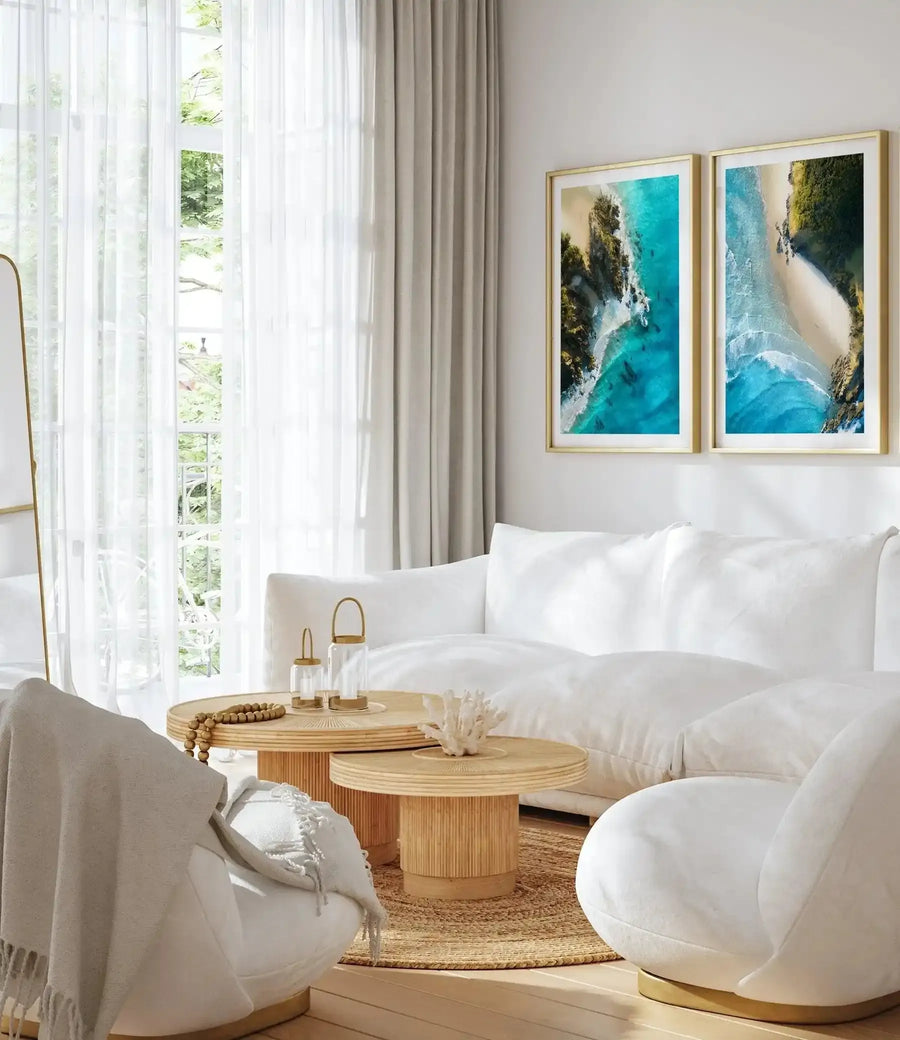 Two framed art prints displayed side-by-side above a white sofa in a bright, minimalist living room. Each print features an aerial photograph of a tropical beach with turquoise water, white sand, and lush green cliffs. The room includes two round wooden coffee tables, a woven rug, and a white armchair with a light beige throw blanket.