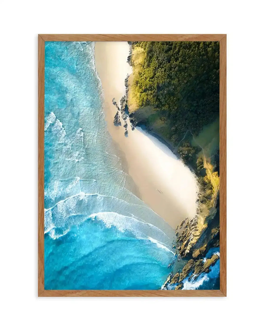 Framed aerial photography art print of a coastal beach scene with natural wood frame and white border. The composition features a long stretch of golden sand bordered by turquoise ocean with white waves breaking, and a lush green forest meeting the sand and rocky outcrops.