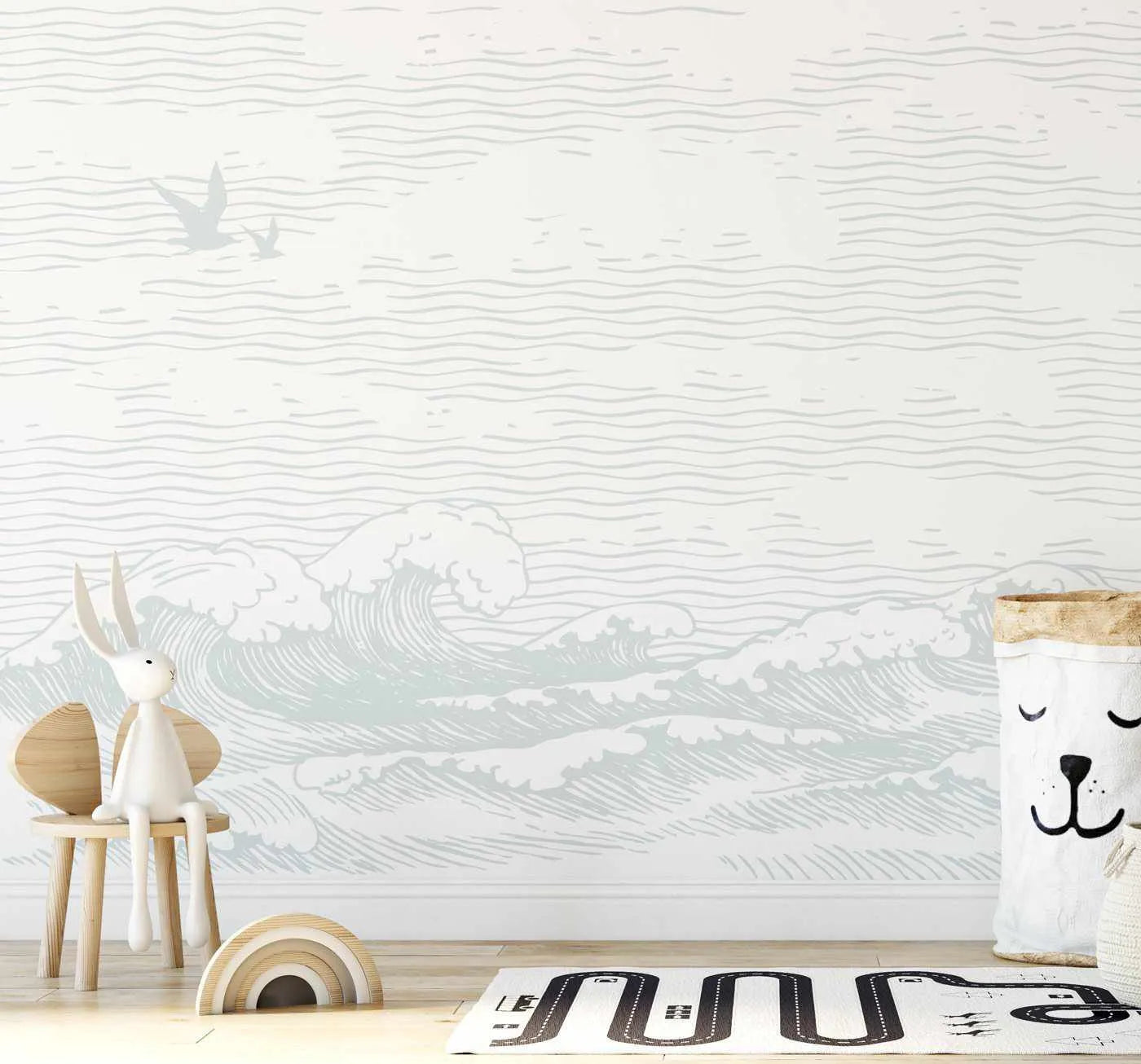 Nursery:
"Peel and stick wallpaper mural featuring a hand-drawn illustration of ocean waves in a soft, muted sage green on a white background, with two seagulls flying in the top left. The mural is displayed on a wall in a child's room with a light wood floor, a white bunny plush toy sitting on a small wooden chair, a wooden rainbow toy, and a white fabric storage basket with a bear face design."