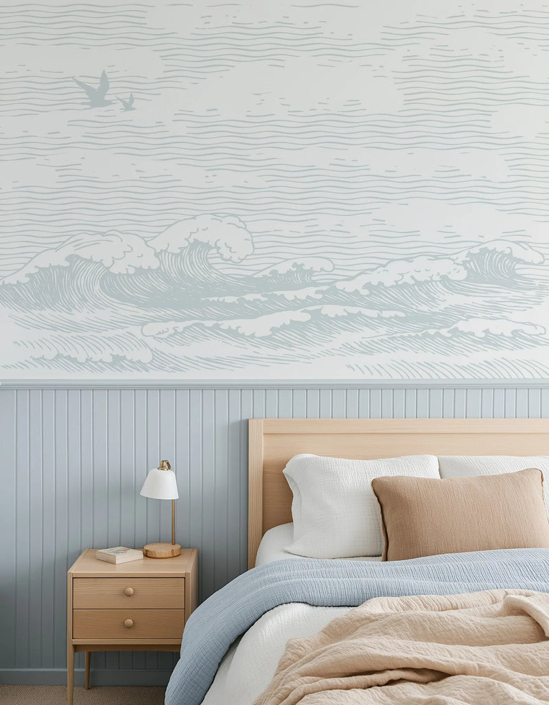 Byron Waves Wallpaper Mural installed on bedroom wall above pale blue beadboard wainscoting. The mural features detailed line-drawn ocean waves in soft sage green and white tones, with birds flying in the upper left corner, rendered in a traditional engraving style. The design creates a serene coastal atmosphere above a wooden bed with light natural wood frame, paired with white and tan bedding and a wooden nightstand with brass hardware and white table lamp.