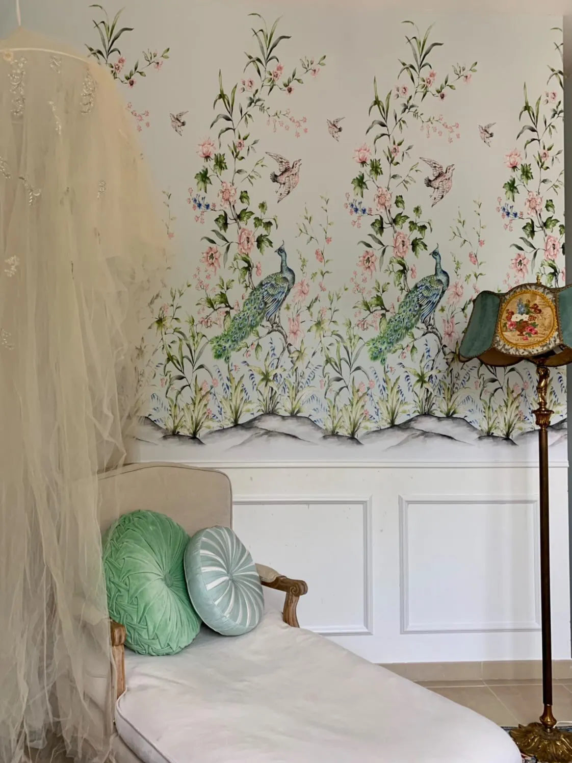 Lifestyle shot of a room featuring Chinoiserie Luxe I wallpaper with a repeating pattern of peacocks and birds amidst flowering branches. The wallpaper has a light background with delicate pink blossoms, green leaves, and blue accents. A white chaise lounge with two round pillows, one green and one light blue with white stripes, is positioned in front of the wallpaper. A sheer white canopy hangs to the left, and a vintage-style floor lamp with a patterned shade stands to the right.