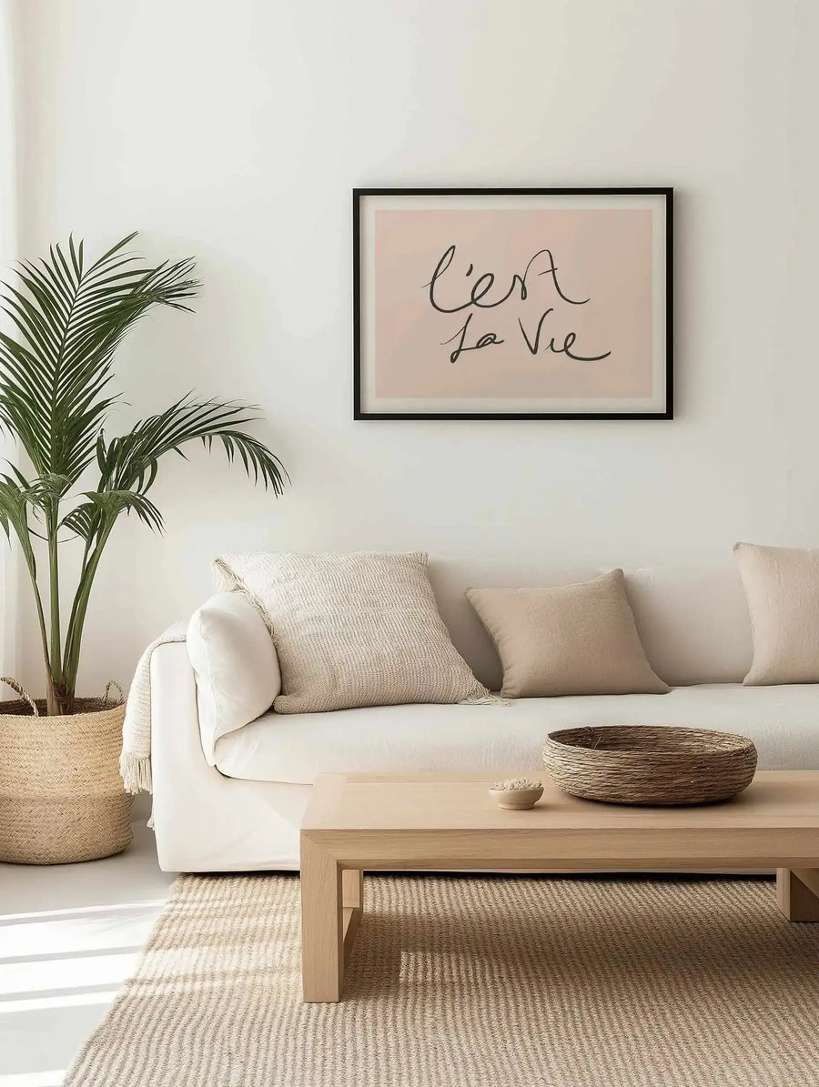 C'est La Vie 2.0 Art Print displayed in a black frame on a cream-colored wall above a cream linen sofa. The print features handwritten script text reading "c'est la vie" in dark gray ink on a soft blush pink background with white border. The artwork is shown in a styled living room with a potted palm plant in a woven basket, cream throw blanket, neutral cushions, light wood coffee table with woven basket, and natural fiber area rug.